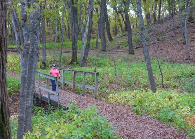 A person wearing a pink shirt and cap stands on a wooden bridge along a leaf-covered hiking trail, surrounded by green and brown foliage in Camden State Park’s forest during autumn.
