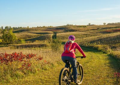 A person wearing a pink helmet and red jacket bikes along a grassy mountain bike trail through rolling hills and tall grass at Camden State Park under a clear blue sky.