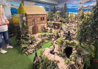 A woman stands next to a large, detailed diorama, featuring miniature figures, a rustic building, trees, rocks, and painted mountains—at Cinnamon Hen in Tracy, MN.