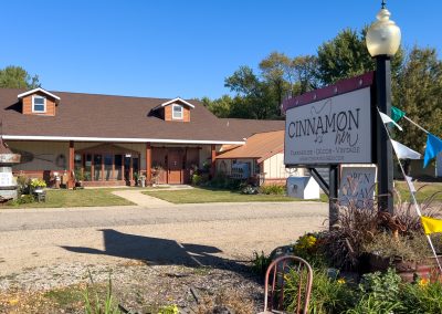 A rustic shop called Cinnamon Hen with a brown roof and large front porch sits along a rural road. A sign featuring a cinnamon hen and colorful pennant flags are in front, surrounded by plants, under a clear blue sky.