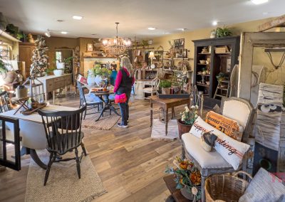 A person browses a cozy shop filled with rustic home decor, wooden furniture, plants, and autumn-themed decorations from Cinnamon Hen. Warm lighting and wood floors create a welcoming atmosphere.
