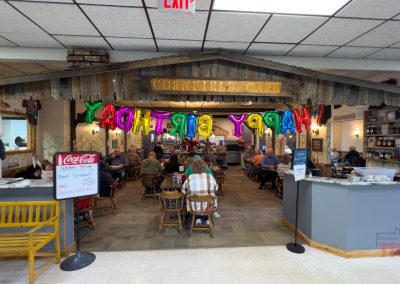 A rustic café with wooden tables and chairs is decorated with large, colorful HAPPY BIRTHDAY balloons. People are dining inside, and a counter with menus and items for sale is visible on the right.