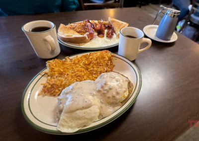 Two plates of breakfast sit on a wooden table: one with eggs covered in gravy, hash browns, and toast, and the other with eggs, bacon, hash browns, and toast. Two mugs of coffee and a metal syrup pitcher are also present.