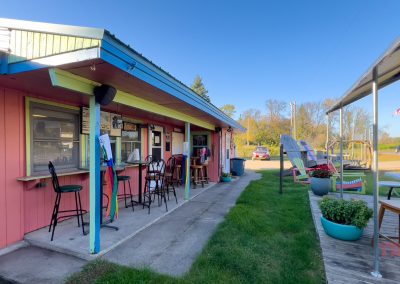 A small pink diner with a turquoise roof, outdoor seating, colorful chairs, and an American flag. Cars are parked in the background, and the area is bright and sunny with green grass and trees.