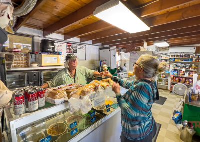 A woman at a small shop counter hands money to a smiling cashier. The counter displays bread and food items, while shelves in the background are stocked with snacks and groceries. The shop has a cozy, rustic feel with wooden beams.