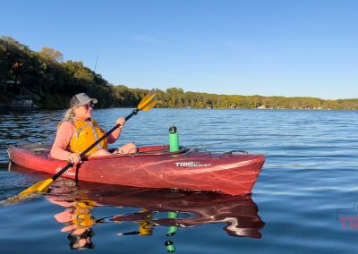 A person wearing a life jacket and hat paddles a red kayak on a calm lake, with trees and houses along the distant shoreline under a clear blue sky. A water bottle is secured on the kayak.