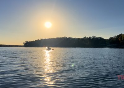 A person is paddling a kayak on a calm lake at sunset, with sunlight reflecting on the water and trees lining the shore.