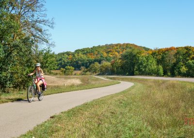 A person wearing a helmet rides a bicycle along the Heart of the Lakes Bike Trail, passing through grassy fields and trees with early autumn foliage under a clear blue sky.