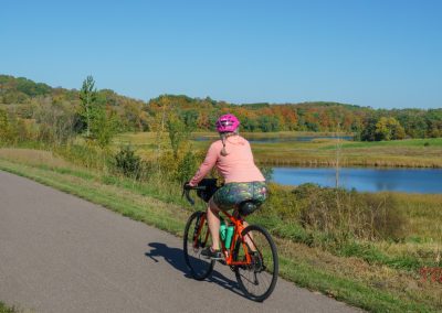 A person wearing a pink helmet and floral shorts rides a bicycle on the Heart of the Lakes Bike Trail beside a pond, with green trees and a blue sky in the background.
