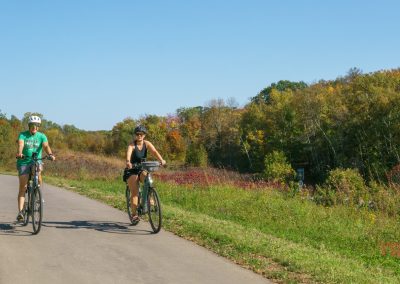 Two people ride bikes on a paved trail through a scenic area with green grass and trees showing early autumn colors under a clear blue sky along the Heart of the Lakes Bike Trail at Maplewood State Park.