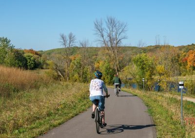 Two people ride bicycles on the Heart of the Lakes Bike Trail, winding through a scenic, tree-filled landscape with autumn foliage in Maplewood State Park under a clear blue sky.