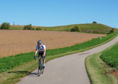 A cyclist wearing a helmet and sunglasses rides along the Heart of the Lakes Bike Trail, surrounded by fields and gentle hills under a clear blue sky.