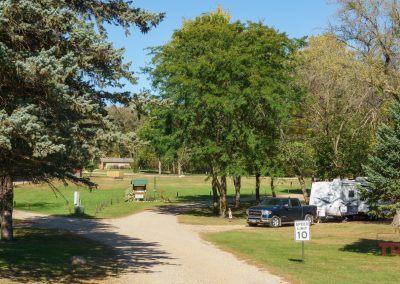 A gravel road curves through Garvin Park Campground, a sunny, green retreat with trees, grass, a parked truck, and a white RV. A “Speed Limit 10” sign is visible near the RV. The peaceful scene feels welcoming and inviting.
