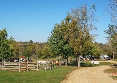 A dirt road curves through Garvin Park’s rural area, where trees, wooden fences, horses, and trailers set the scene for a relaxing day of horseback riding under a clear blue sky.