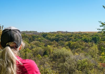 A person with long blonde hair, wearing a black mesh cap and a red shirt, stands on a viewpoint along the Garvin Park Trails, looking out over a forested landscape under a clear blue sky, enjoying a day of hiking.