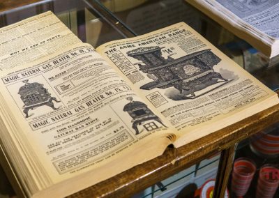 An open vintage catalog on display at the Lyon Co History Museum shows illustrations and ads for antique stoves and heaters, including The Acme American Range, with old-fashioned fonts and detailed product drawings.