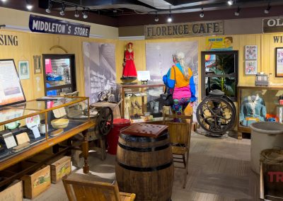 A museum exhibit at Lyon Co History Museum Exhibits features vintage items like display cases, a barrel, washing machine, and mannequins in period clothing. Signs read “Middleton’s Store,” “Florence Cafe,” and “Wish Book.” A visitor observes the displays.