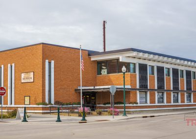 A modern brick building with large windows houses the Lyon Co History Museum. An American flag flies outside, and street lamps line the sidewalks at the street corner on a cloudy day.