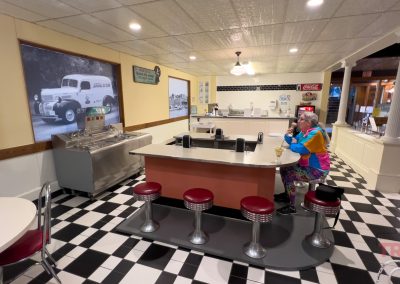 A person in colorful clothes sits alone at the counter of a retro-style diner with black-and-white checkered floors, red stools, and vintage decor reminiscent of an Ice Cream Parlor at the Lyon Co History Museum.