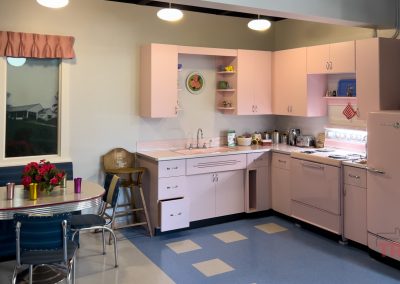 A retro 1950s-style Lyon Co History Museum Kitchen with pink cabinets, stove, and fridge, a round table with blue chairs, a vase of red flowers, and pastel décor creates a vintage, cozy atmosphere.
