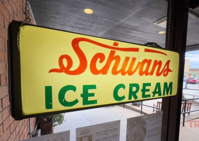 A vintage illuminated Schwan’s Sign glows in a storefront window at the Lyon Co History Museum. Red and green letters stand out near a brick wall, with a lively street scene visible outside.