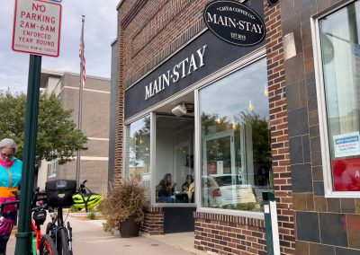 A person in colorful clothing stands next to a parked bicycle outside Main-Stay Coffee, with people visible inside through large windows. A No Parking sign is posted on the sidewalk.