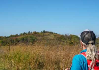 A person with long gray hair, wearing a mesh cap, blue shirt, and red backpack, stands in tall grass at Maplewood State Park, hiking while gazing toward a distant green hill under a clear blue sky.