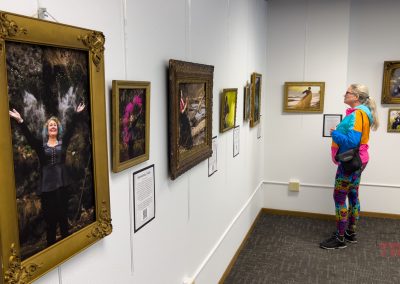 A woman in colorful clothes studies framed photographs on a white gallery wall at the Marshall Area Fine Arts exhibit, including portraits and nature scenes. One large photo shows a smiling woman with arms raised in front of a waterfall.