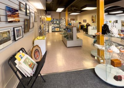 An art gallery interior at Marshall Area Fine Arts with framed artwork on the walls, a jewelry display on glass shelves, and people working at desks in the background. The space features light-colored walls and a polished tile floor.