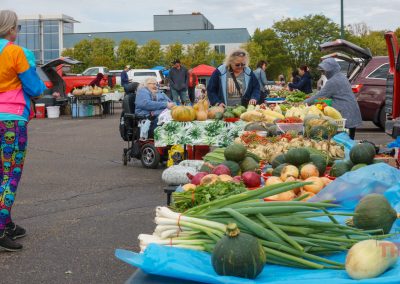 Customers browse tables of fresh vegetables and produce at the Marshall Farmers Market. A woman wearing colorful leggings stands nearby as people shop or sit behind tables displaying pumpkins, onions, squash, and other vibrant vegetables.