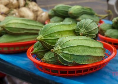 Several ridged green vegetables, likely angled luffa gourds, are displayed in red plastic baskets on a blue surface at the Marshall Farmers Market. Other fresh produce can be seen in the background.