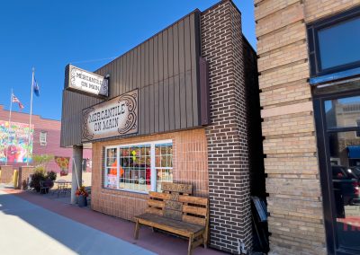 A small brick and brown metal storefront with a sign reading Mercantile on Main. A wooden bench sits outside the window, and American flags are visible on the street. The sky is clear and blue.