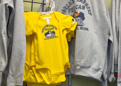 A yellow baby onesie with Worlds Largest Pelican, Pelican Rapids, Minn. printed on it hangs among gray sweatshirts with the same design.