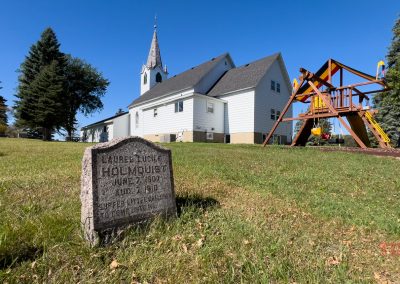 A gravestone sits on a grassy hill in front of a white church with a tall steeple. To the right, a colorful playground with swings and a slide is visible under a clear blue sky.