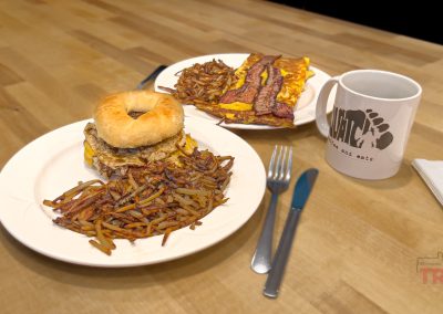 A breakfast at Squatch Coffee And Eats: a bagel sandwich with eggs and hash browns, another plate with an omelet and hash browns, plus a mug, fork, knife, and napkin on a wooden table.