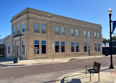 A two-story beige stone building on a street corner with large windows and glass blocks near the roofline. A bench and streetlamp are in the foreground, while blue banners for Squatch Coffee And Eats decorate the area on a sunny day.
