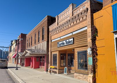 A small-town main street with historic brick buildings, in Tracy, MN, including Tattle Tales, a charming local shop. The street is quiet under clear blue skies, with industrial silos visible in the background.