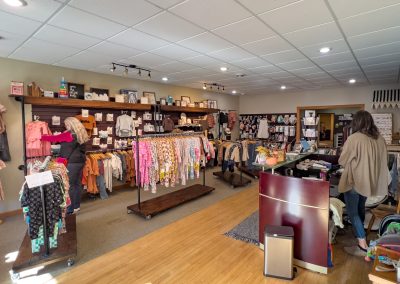 Tattle Tales, a boutique clothing store in Tracy, MN, features wooden shelves and racks displaying colorful children's clothes. Two women are shopping while the cozy, brightly lit atmosphere surrounds the checkout counter in the foreground.
