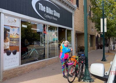 A person in colorful cycling gear stands next to a bicycle on the sidewalk outside The Bike Shop, Marshall, which features large windows. Trees and parked cars line the street.