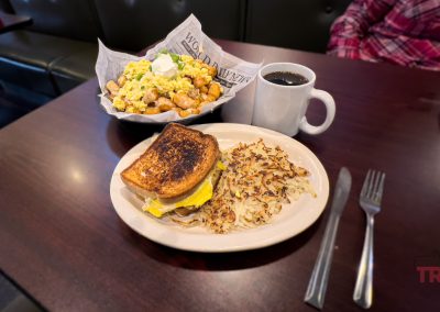 A breakfast table at The Wooden Nickel with a plate of hash browns and a sandwich, a bowl of scrambled eggs with tater tots, a mug of coffee, utensils, and a person in a red plaid shirt seated at the booth.