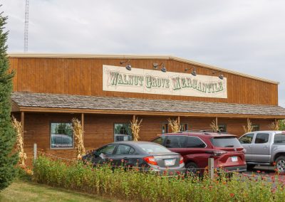 A rustic wooden building with a sign reading Walnut Grove Mercantile above the entrance. Several cars are parked in front, and corn stalks decorate the porch of Walnut Grove Mercantile. Trees and greenery are visible nearby.