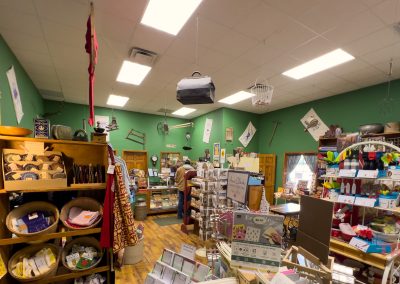 A small, cozy gift shop with green walls, shelves filled with various items, greeting cards, baskets, and souvenirs. Two people stand near the counter at the back of Walnut Grove Mercantile, and the ceiling is decorated with local memorabilia.