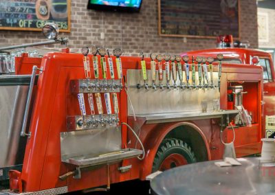 A vintage red fire truck is converted into a bar with multiple beer taps mounted on its side, set indoors against a brick wall with chalkboard menus and glasses visible in the background.