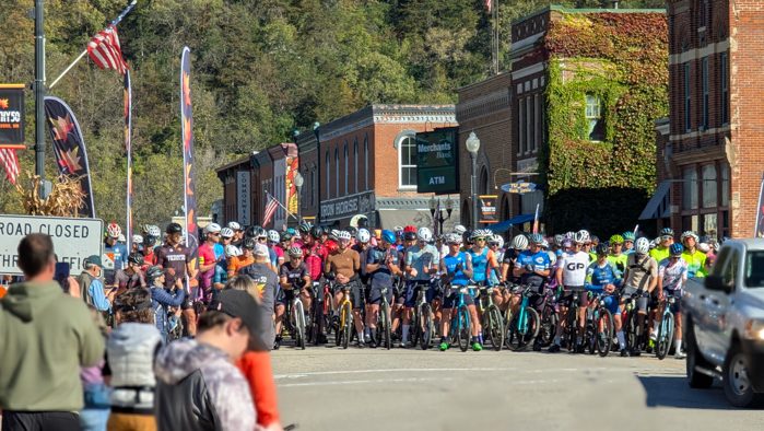 Filthy 50_Lanesboro Chamber A large group of cyclists wearing helmets and colorful jerseys line up at the starting line of the Filthy Fifty Gravel Race in Lanesboro, MN, surrounded by brick buildings and onlookers. The street is blocked to vehicles.