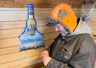 A man in a winter hat and coat stands inside a wooden cabin at Rainbow Resort in Waubun, MN, looking down at something in his hands. Next to him is a wall sign shaped like a bottle of Michelob Ultra with text that reads Its Pure Cold.