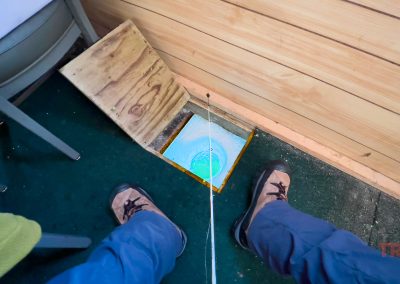 A person ice fishing indoors at Rainbow Resort in Waubun, MN sits by a small wooden trapdoor in the floor, open to reveal an ice hole with a fishing line dropped in. Their boots and legs are visible beside the cozy setup.