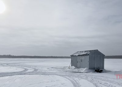 A small metal ice fishing shack sits on a snow-covered frozen lake near Rainbow Resort in Waubun, MN. Tire tracks curve beside the shack as sunlight filters through cloudy skies, with distant trees lining the horizon.