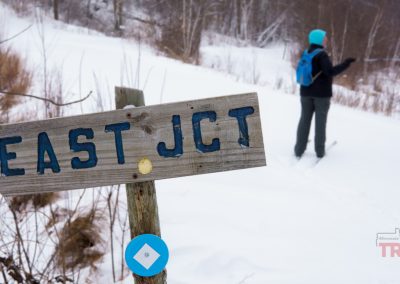 A weathered wooden sign reading EAST JCT stands in the snow at Rainbow Resort in Waubun, MN, with a person in winter clothing and a blue backpack skiing on a snowy trail in the background.