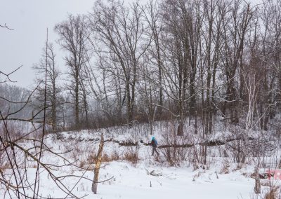 A person in a blue jacket walks through a snowy landscape near Rainbow Resort in Waubun, MN, with leafless trees and bushes in the background on a cloudy winter day.