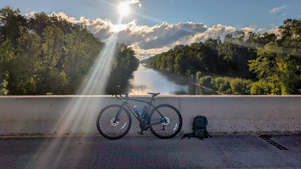 2025 BAM Bike Tour-River Bike A bicycle and a backpack rest against a concrete bridge railing, with sun rays beaming down over a calm river—an inviting scene along Minnesota's BAM Bike Tour, flanked by lush green trees under a partly cloudy sky.